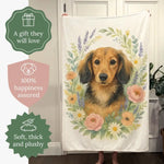 A person holds up the blanket showing a long-haired dachshund in a pastel floral wreath; badges read "A gift they will love", "100% happiness assured" and "Soft, thick and plushy".