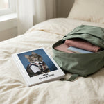 High School Photo Pet Notebook. The white hardcover notebook on rumpled cream bedding beside an open backpack, soft morning light across the duvet.