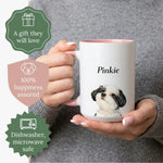 Hands holding a white mug with a pink handle showing a watercolor Shih Tzu named "Pinkie". Badges read "A gift they will love", "100% happiness assured" and "Dishwasher, microwave safe".