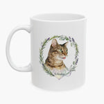 Watercolor portrait of a winking tabby cat inside a rosemary and lavender wreath on a white mug, with the name Whiskey on a heart below.