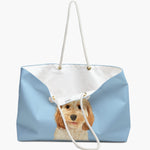The French blue weekender bag standing open, its white cotton lining showing, with the watercolor apricot puppy portrait below.