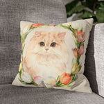 The cream tulip-wreath pillow with the fluffy Persian cat design, propped on a gray woven couch beside a leafy green houseplant.