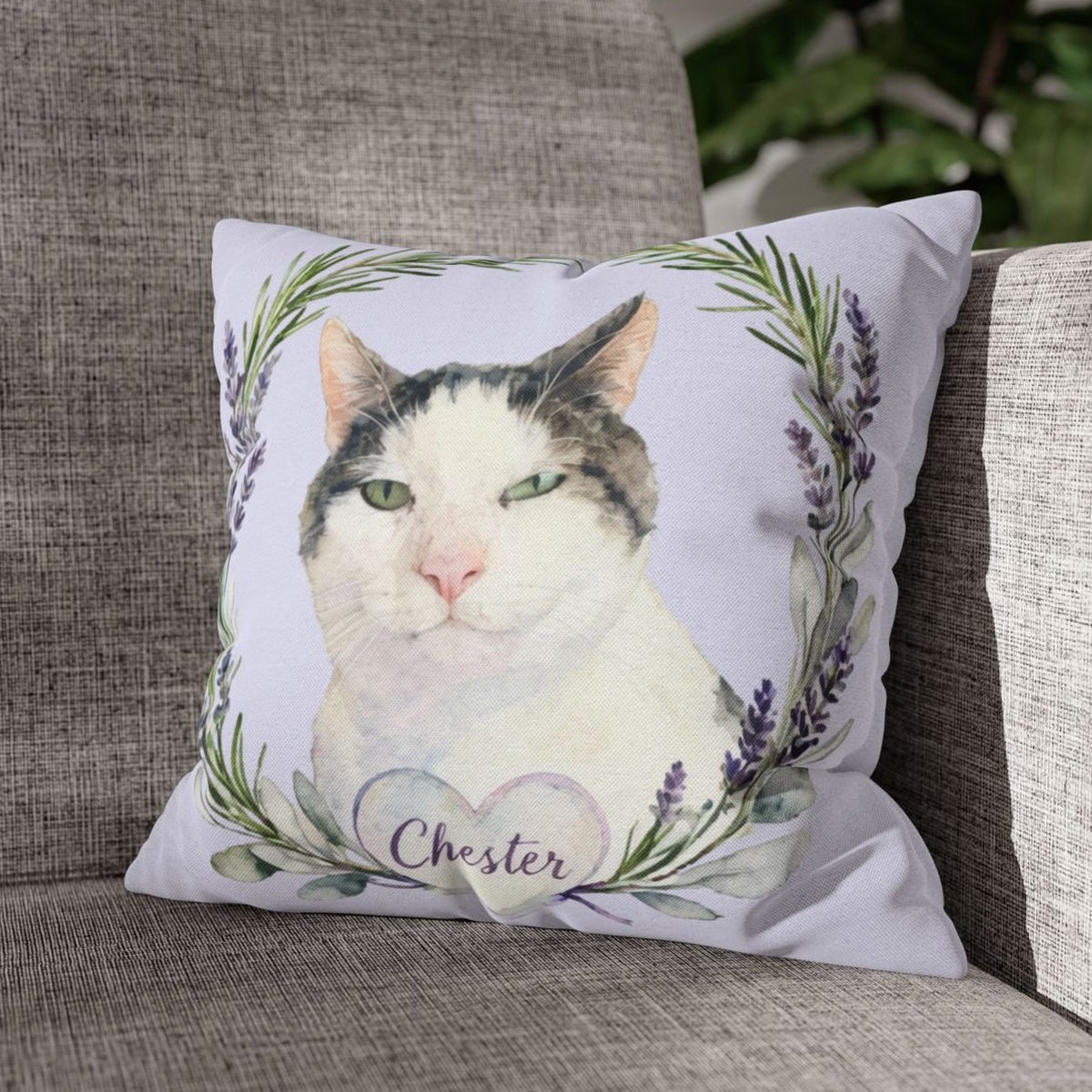 The pale lilac lavender-wreath pillow with the gray and white cat design, propped on a gray woven couch beside a green houseplant.
