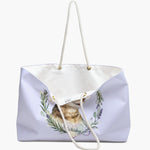The lilac herb-wreath weekender bag standing open, white lining and care label showing, with the tabby cat and "Whiskey" heart below.