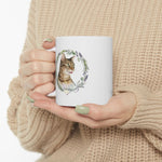 Hands in a beige knit sweater holding the white mug, showing the tabby cat Whiskey framed by a rosemary and lavender wreath.