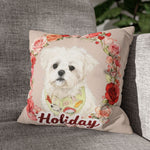 The beige rose-wreath pillow showing a white Maltese in a patterned bib and the name "Holiday", propped on a gray woven couch.