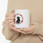 Hands in a beige knit sweater holding the white mug, showing a black cat named Popo inside a wreath of roses and hearts.