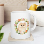The mug on a kitchen counter beside a bowl of lemons and an avocado, showing the cream Persian cat in a watercolor tulip wreath.