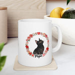 The mug on a kitchen counter beside a bowl of lemons and an avocado, showing the black cat inside a wreath of roses and hearts.