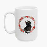 Black cat looking upward inside a wreath of roses and hearts, printed on the taller white mug with the name Popo below.