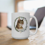 The larger mug on a wooden desk in front of a laptop, showing the tabby cat Whiskey inside a rosemary and lavender wreath.