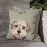 The sage green pillow with the terrier "JOJO" design, leaning in the corner of a gray woven couch next to a leafy green houseplant.