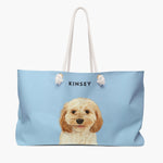 Watercolor portrait of an apricot cavapoo on a French blue weekender bag with white rope handles, with the name KINSEY printed above.